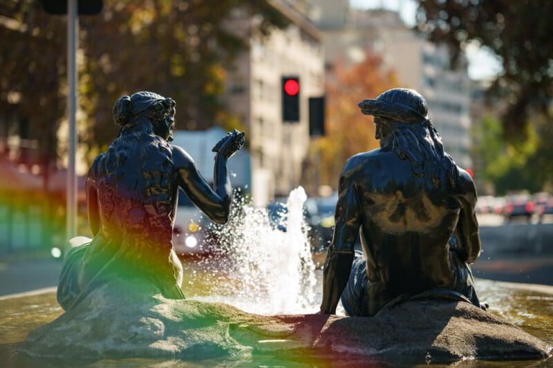 Two dark statues of seated figures face a small fountain on a city street, with a rainbow lens flare across the foreground and blurred buildings and traffic lights in the background.