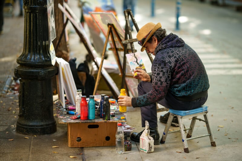 A street artist wearing a hat and patterned hoodie sits on a stool, painting at an outdoor easel with colorful bottles of paint on a low table beside him. The scene takes place on a city sidewalk.