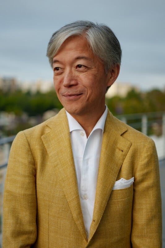 A man with gray hair wearing a mustard yellow blazer, white dress shirt, and white pocket square stands outdoors, smiling softly with a blurred cityscape in the background.