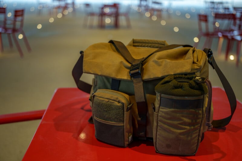 A brown messenger bag with multiple pockets sits on a red table. In the blurred background, several red chairs are spaced apart in a large, open indoor area.