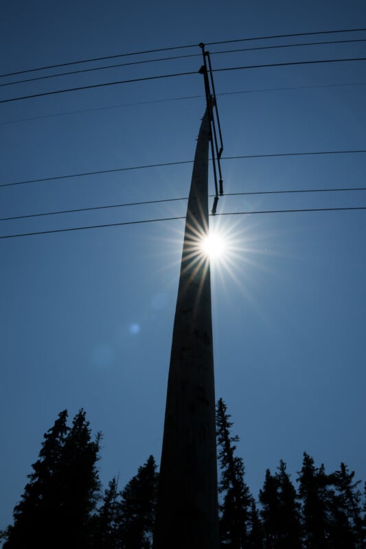 A utility pole with power lines is silhouetted against a clear blue sky, with the sun shining brightly behind it. Pine trees are visible at the bottom of the image.
