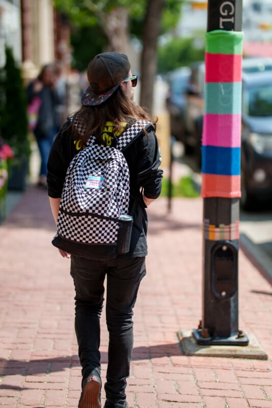 A person with long hair, a cap, and a checkered backpack walks on a brick sidewalk next to a pole wrapped in colorful yarn. Cars and trees line the street.