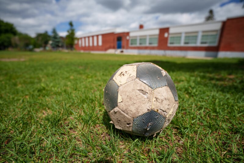 A worn and weathered soccer ball sits on green grass, with a red-brick school building and blue sky with clouds in the background.