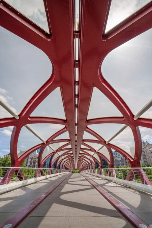 A low-angle view of a modern red pedestrian bridge with a geometric, tubular structure and glass roof, stretching over a river toward city buildings under a partly cloudy sky.