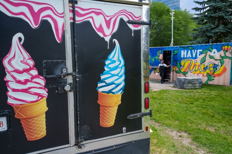 An ice cream truck with illustrated cones is parked on grass near a colorful mural that says "Have a Nice Day," with a person standing nearby. Trees and buildings are visible in the background.