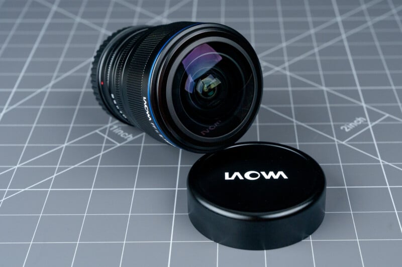 A Laowa camera lens rests on a gray grid-patterned surface next to its detached lens cap, which displays the brand name "LAOWA" in white letters.