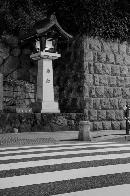 A black and white image of a Japanese stone lantern with inscriptions, mounted on a pedestal beside a cobblestone wall. A zebra crossing is in the foreground, and surrounding greenery partially covers the wall.