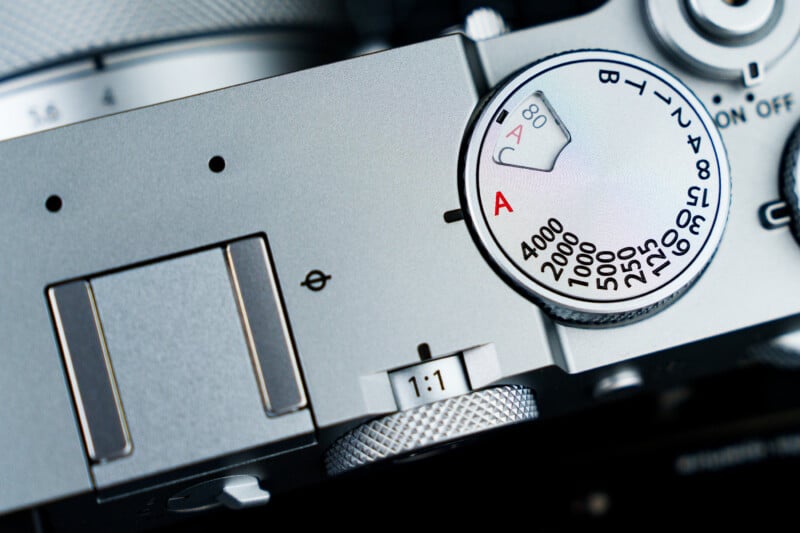 Close-up of a silver camera's top panel featuring a mode dial with settings in black and red text. The dial includes numbers indicating shutter speeds from 30 to 4000. Adjacent to it are various buttons and controls on a gray metallic surface.