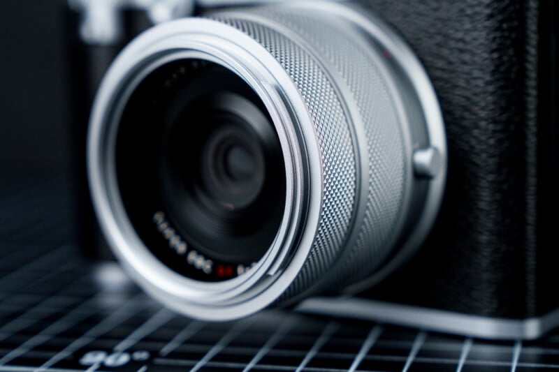 Close-up of a vintage-style camera lens with a textured metallic ring, positioned on a surface marked with a black-and-white grid pattern. The focus is on the intricate details of the lens design.