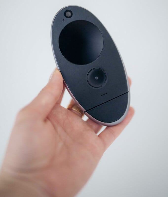 A hand holding a small, oval-shaped black electronic device with a camera lens and a circular touchpad on its surface against a plain light background.