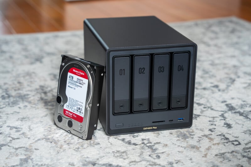 A black 4-bay NAS enclosure labeled 01 to 04 stands on a gray carpet, with a 1TB Western Digital Red hard drive placed upright next to it.