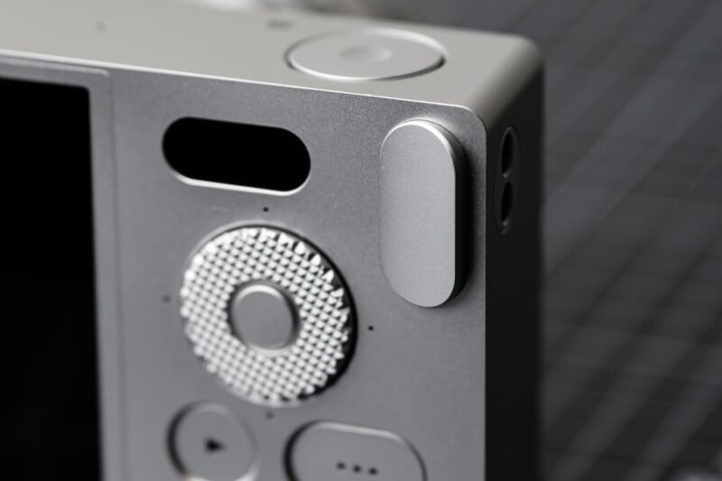 Close-up of a silver digital camera, showcasing its textured control dial, buttons, and viewfinder. The surface has a sleek, metallic finish, and the background is a blurred gray grid pattern.