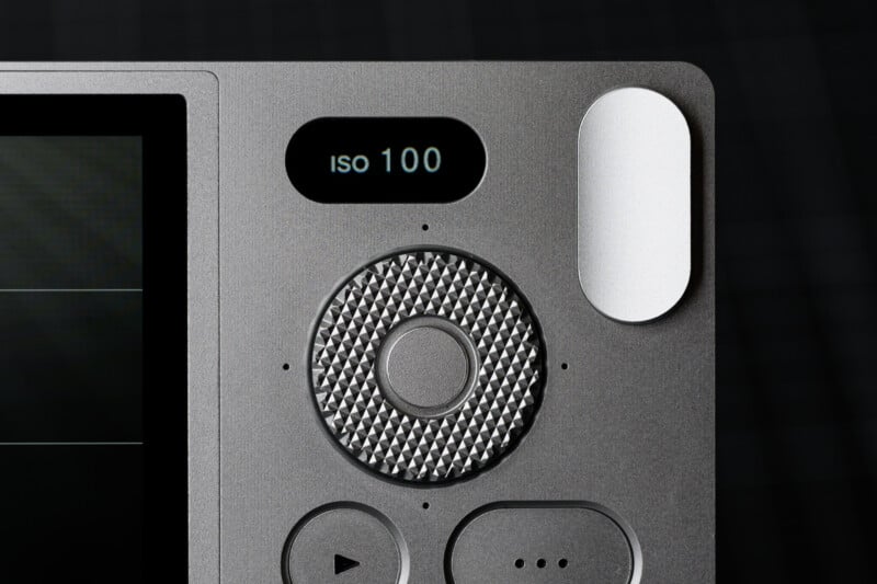 Close-up of a grey camera interface showing a textured control dial, display reading "ISO 100," and minimalist function buttons, set against a dark, blurred background.
