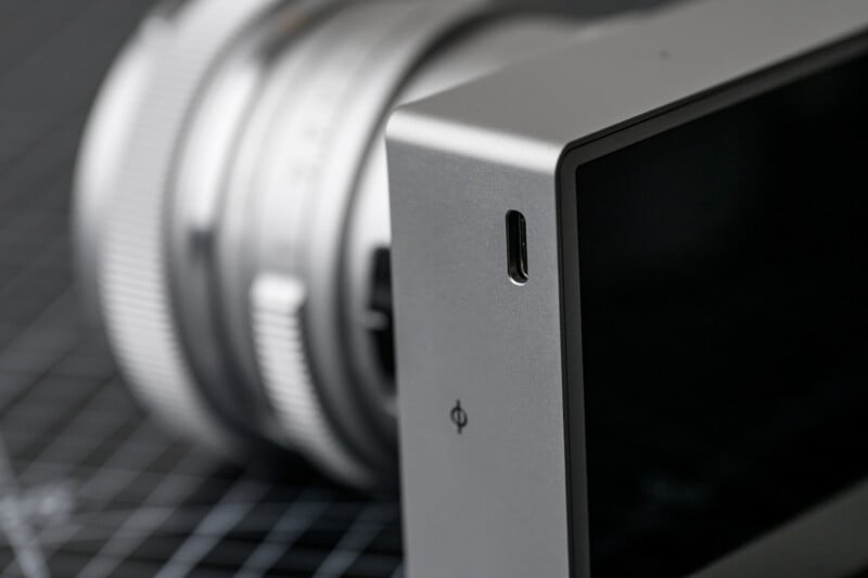 Close-up of a digital camera, focusing on the edge of its screen and body. The background shows the camera lens with silver detailing, all set against a black grid-patterned surface.