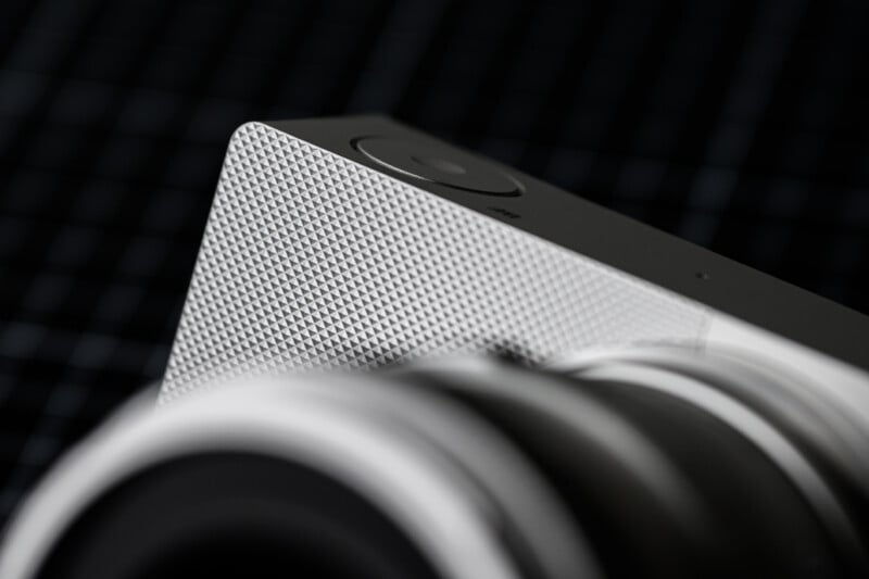 Close-up of a stylish camera, focusing on its textured silver surface and a circular button. The sleek design highlights the geometric pattern, with the lens slightly blurred in the foreground against a dark, grid-like background.