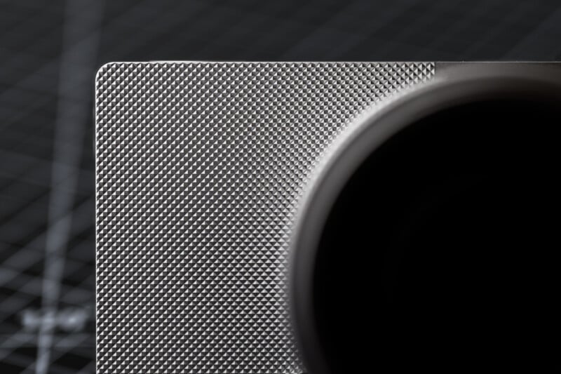 Close-up of a textured metal surface with a grid pattern, featuring a large circular black shape partially covering the right side. The background shows a blurred grid design, suggesting a technical or industrial context.