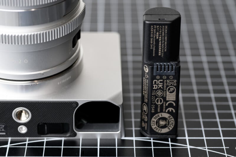Close-up of a camera mount and a battery on a grid-patterned surface. The silver camera mount has a textured section, while the black battery stands upright with visible labels and symbols.