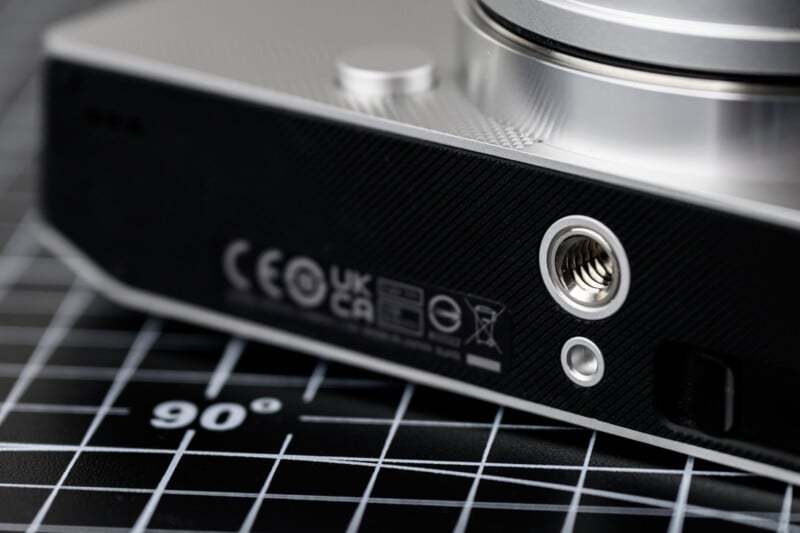 Close-up of a camera placed on a black grid mat, showcasing the bottom part with various certification logos, a tripod mount, and textured grip. The mat has white grid lines and a 90-degree angle marking.
