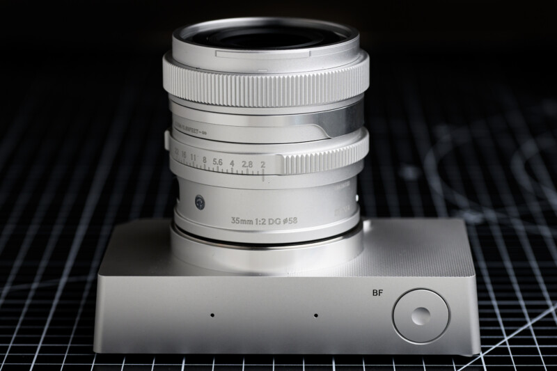 A silver camera lens is mounted on a matching metallic stand. The lens has engraved numbers indicating focal length, and the stand features two small holes and a dial labeled "BF." The background is a black and white grid surface.