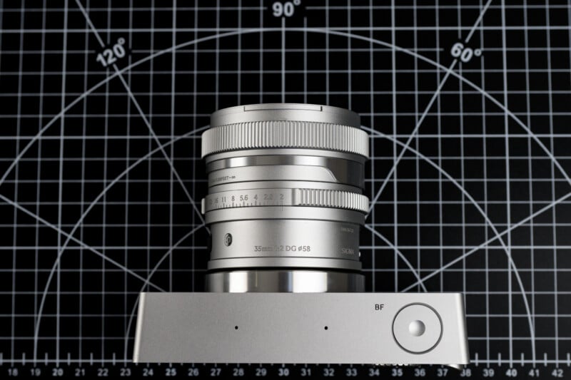 A silver camera lens is placed upright on a graphing grid background with markings in degrees. The lens rests on a rectangular metallic base with a circular button labeled "BF.