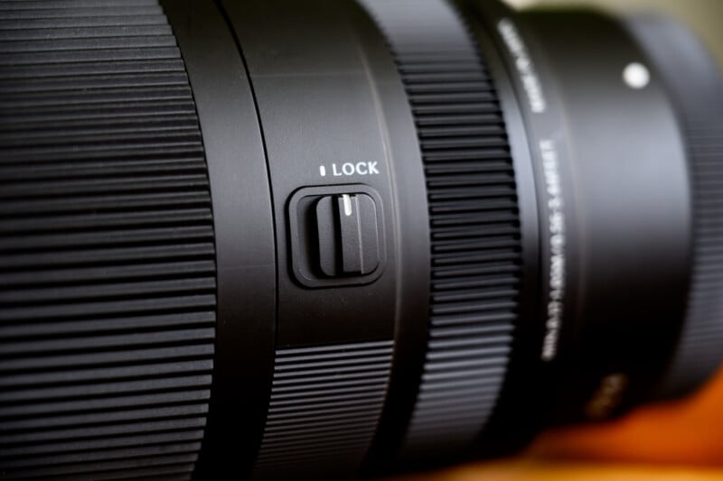 Close-up of a black camera lens with a visible lock switch on its body. The lens features textured ridges for grip and has white text near the rim. The focus is on the lock switch, with a blurred background.