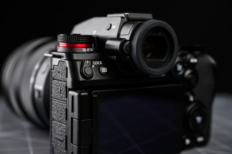 A close-up view of the back and side of a black digital camera, showing buttons, a textured grip, and part of the lens, with a dark, out-of-focus background.