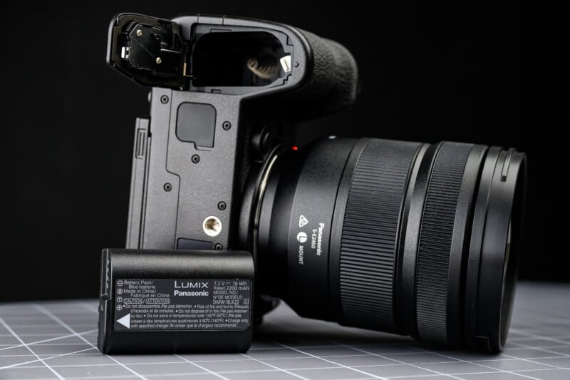 A Panasonic Lumix camera with its battery compartment open sits on a gray grid-patterned surface. The battery is removed and placed in front of the camera, showing its label and specifications.