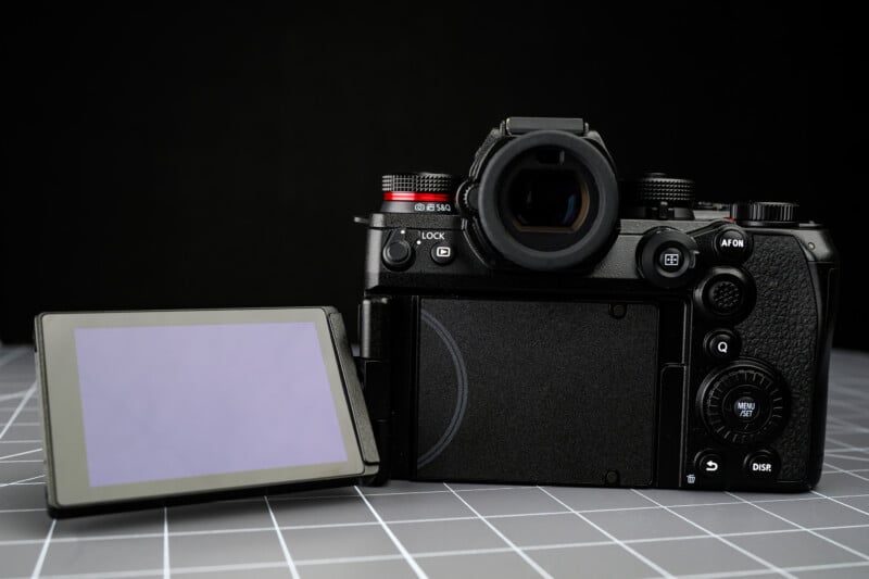 A digital camera with its rear LCD screen flipped out to the left sits on a gray, grid-patterned surface against a black background. Buttons and controls are visible on the back of the camera.