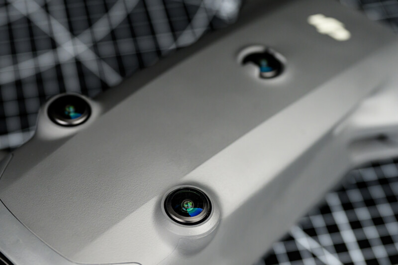 Close-up of a gray drone showing three camera sensors on its body, placed on a black and white grid-patterned surface.