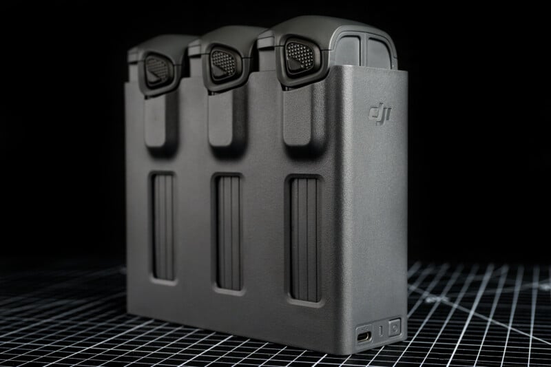 A close-up of a DJI battery charging hub holding three drone batteries, placed on a grid-patterned surface with a black background.