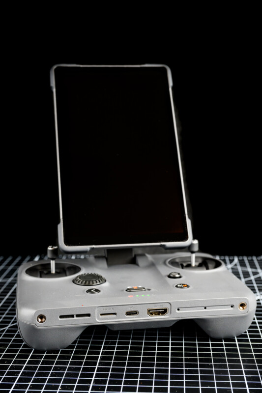A gray remote controller with a large black screen attached, possibly for a drone, is placed on a grid-patterned surface with a black background.