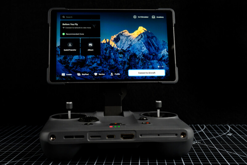 A close-up of a drone remote controller with a screen displaying a mountain landscape and flight settings against a black background. The controller is placed on a grid-patterned surface.