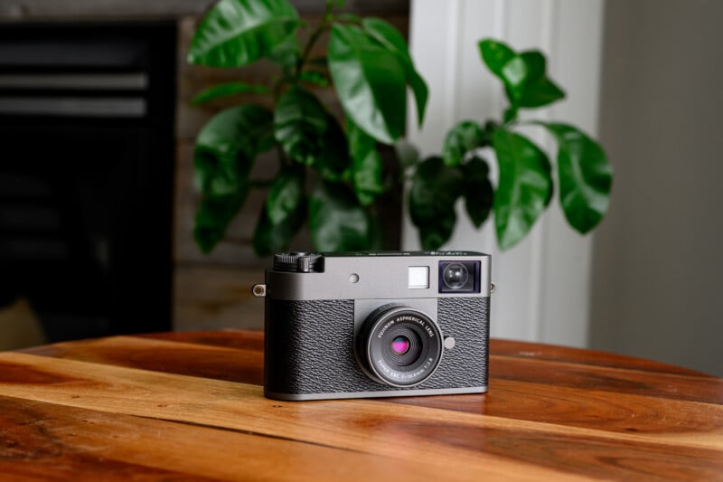A black and silver vintage-style camera sits on a wooden table with a green leafy plant and white wall in the background.