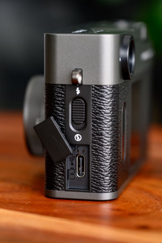 Close-up of the side of a black camera showing a textured grip, a covered USB port, flash switch, and other buttons, placed on a wooden surface.