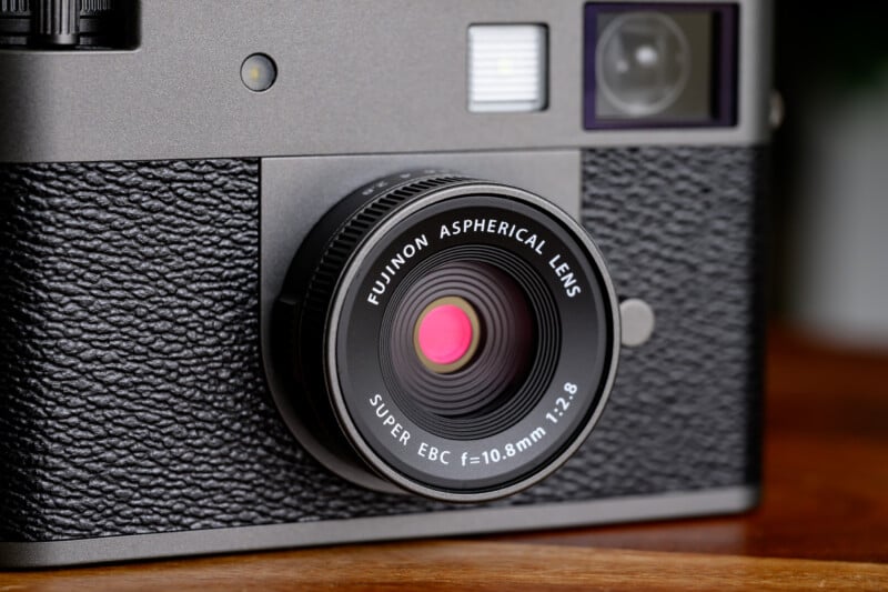 Close-up of a camera showing a Fujinon Aspherical Lens, Super EBC f=10.8mm 1:2.8, mounted on a textured black and silver camera body, with parts of the viewfinder and flash visible.