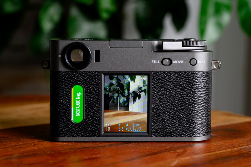 A digital camera with a textured black body is sitting on a wooden surface. Its display screen shows a photo preview of green leaves on a tabletop. A green label reads "NOSTALGIC Neg." in white text.