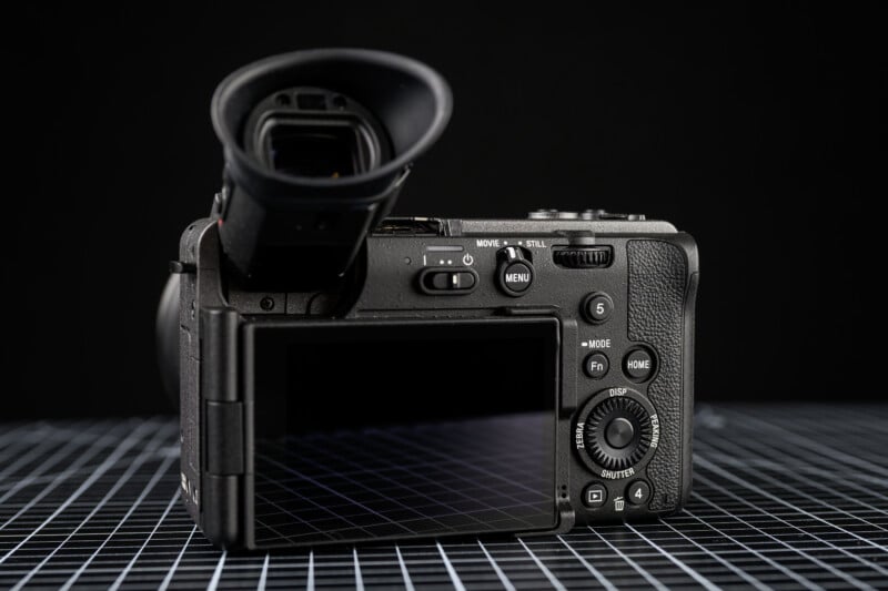 A digital camera with its rear LCD screen open and facing the viewer, showing various buttons and dials, placed on a grid-patterned surface against a black background.