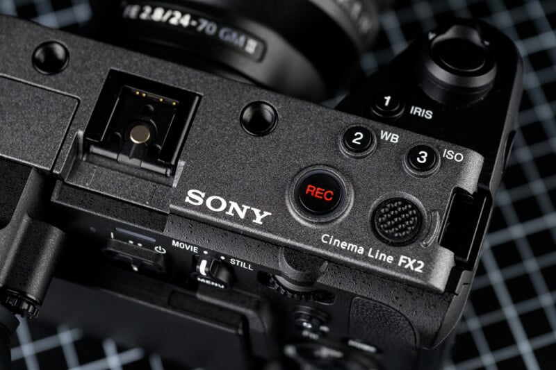 Close-up of the top controls of a black Sony Cinema Line FX2 camera, showing the REC button, dials for iris, white balance, and ISO, with a lens and a grid-patterned surface in the background.