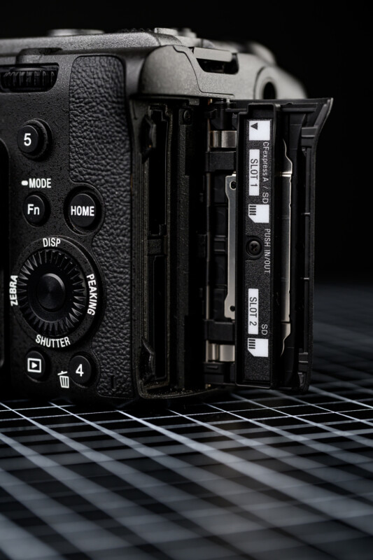 Close-up of a digital camera with its memory card slot door open, showing two SD card slots labeled “Slot 1” and “Slot 2.” The camera rests on a grid-patterned surface.