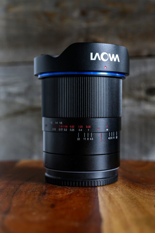 A Laowa camera lens with a petal-shaped lens hood sits upright on a wooden surface, with a rustic, blurred wooden background behind it. The lens features white and red markings and a textured focus ring.
