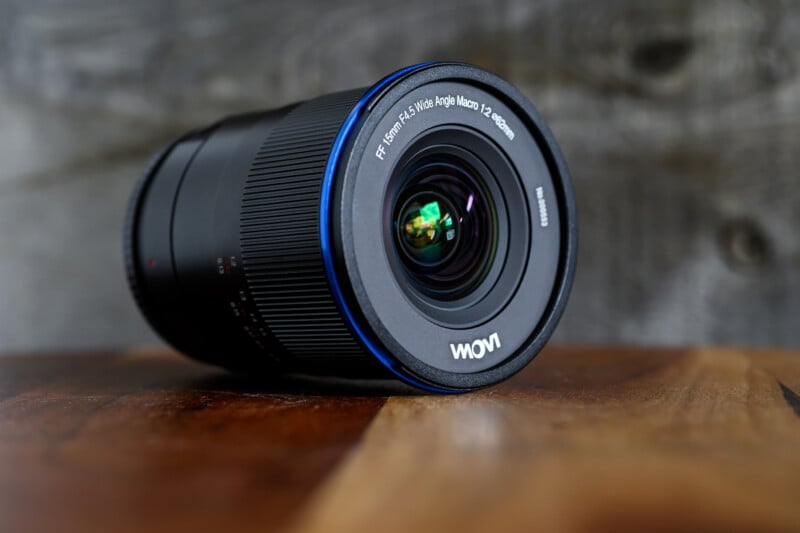 A Laowa camera lens with "FF 15mm f/4.5 W-Dreamer" written on the front, resting on a wooden surface with a blurred, rustic background.