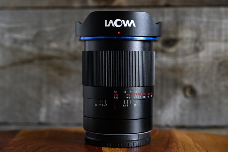 A Laowa camera lens stands upright on a wooden surface with a rustic wooden background. The lens features visible focus and aperture markings in white and red.