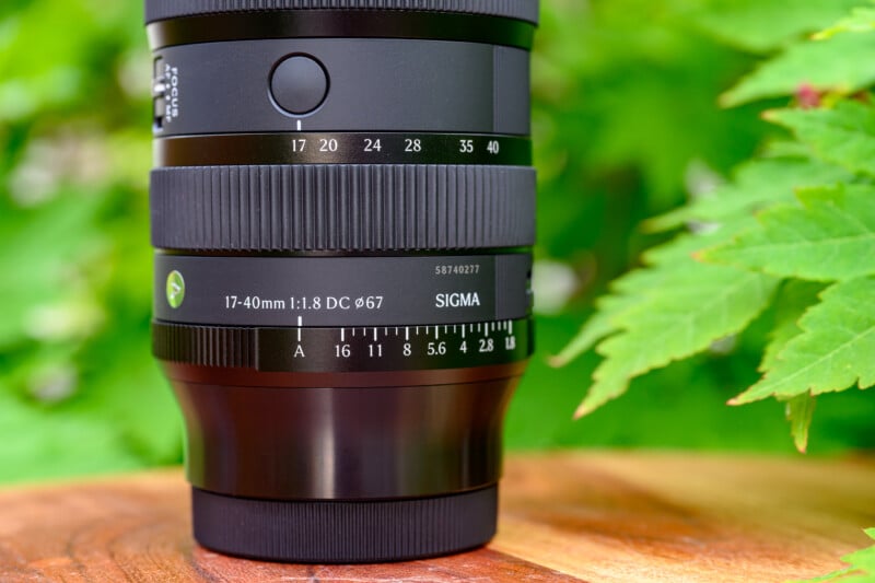 A close-up of a Sigma 17-40mm f/1.8 DC camera lens, resting on a wooden surface with green leaves and blurred foliage in the background.