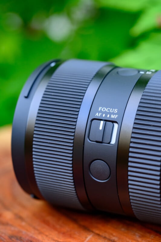 Close-up of a black camera lens with a visible switch labeled “FOCUS AF MF,” resting on a wooden surface with green foliage blurred in the background.