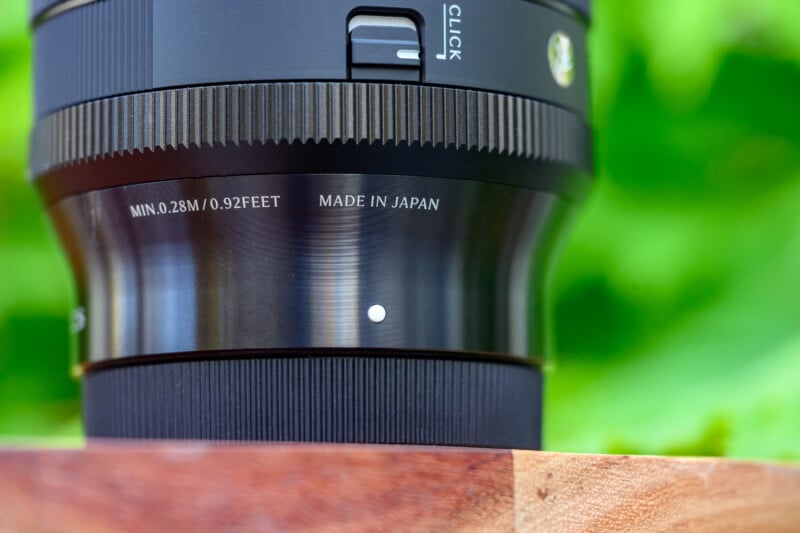 Close-up of a black camera lens with visible text reading "MIN. 0.28M / 0.92FEET" and "MADE IN JAPAN," set against a blurred green background and resting on a wooden surface.