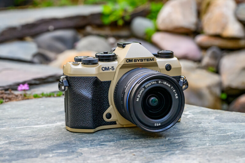A beige and black OM System OM-5 digital camera with a M.Zuiko Digital lens sits on a stone surface outdoors, with blurred rocks and greenery in the background.