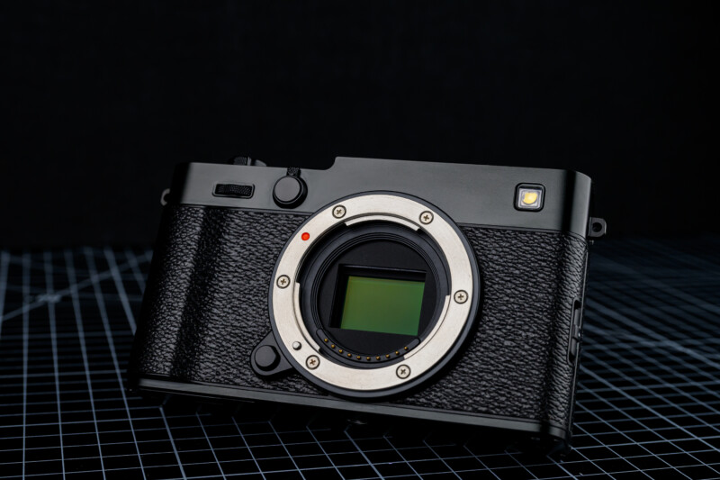 A black mirrorless digital camera body without a lens is placed on a black grid-patterned surface against a dark background. The camera’s sensor and textured grip are clearly visible.