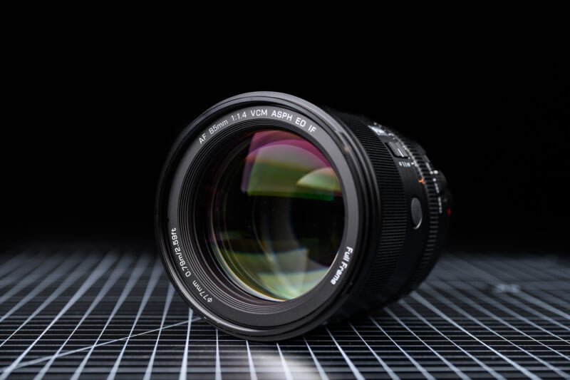 A close-up of a camera lens with "AF 85mm 1:1.4 VCM ASPH ED IF" written on the rim, placed on a black grid-patterned surface, with a black background.
