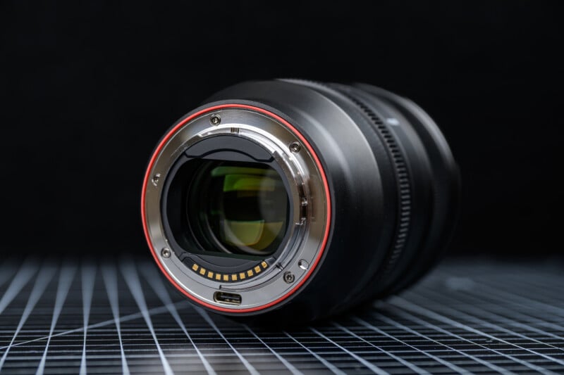 A close-up of a camera lens lying on its side on a grid-patterned surface, showing the metallic mount and electronic contacts, with a black background.