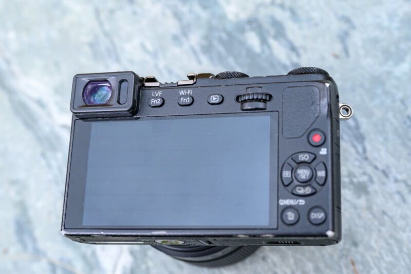 A close-up view of the back of a digital camera with a large screen, various buttons, and a viewfinder, placed on a light gray textured surface.